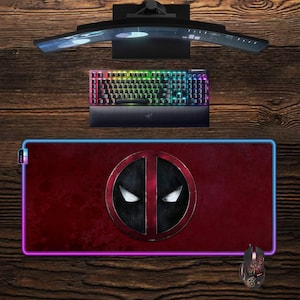 May include: A gaming setup featuring a curved monitor, a backlit keyboard, and a Deadpool-themed mousepad with a matching mouse. The mousepad has a red background with the Deadpool logo and a glowing border.