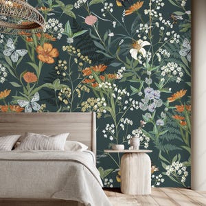 May include: A bedroom with a dark teal floral wallpaper featuring orange, white, and yellow flowers, butterflies, and bees. A wooden bed frame with a light gray blanket and pillows is in the foreground. A small wooden side table is next to the bed.