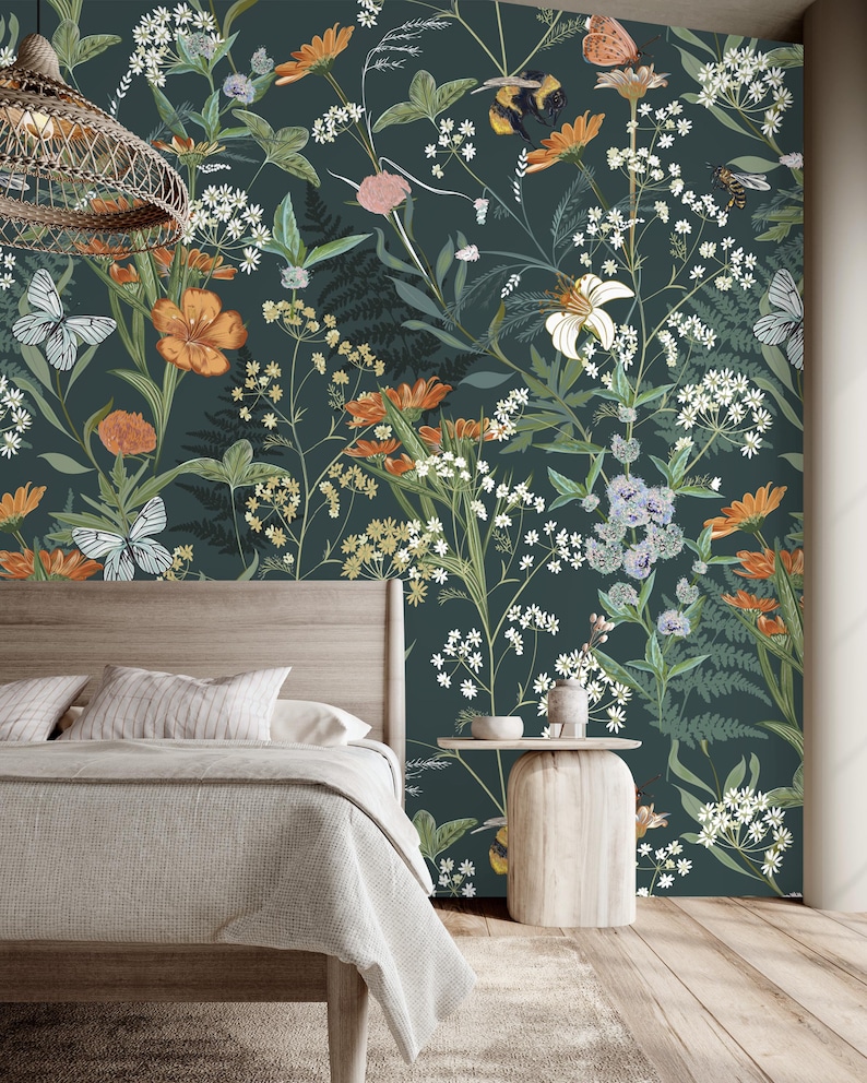 May include: A bedroom with a dark teal floral wallpaper featuring orange, white, and yellow flowers, butterflies, and bees. A wooden bed frame with a light gray blanket and pillows is in the foreground. A small wooden side table is next to the bed.