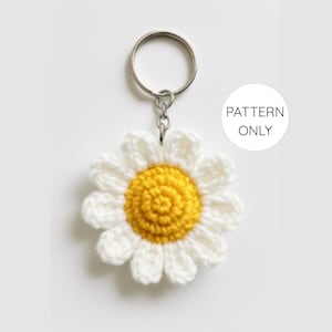 Könnte beinhalten: Ein gehäkelter Schlüsselanhänger in Form einer Gänseblume mit gelber Mitte und weißen Blütenblättern. Der Schlüsselanhänger ist an einem silbernen Metallring befestigt. Das Bild enthält einen weißen Kreis mit dem Text "PATTERN ONLY".