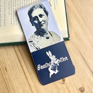 May include: A bookmark featuring a portrait of a woman and the name "Beatrix Potter." The bookmark has a navy blue base with a white illustration of a rabbit and the author's name in white script. The top portion has a portrait.