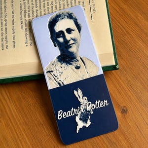 May include: A rectangular bookmark featuring a raised, monochromatic portrait of Beatrix Potter on a light blue background. The lower section is dark blue with a white, raised silhouette of a rabbit and the white text Beatrix Potter in script.