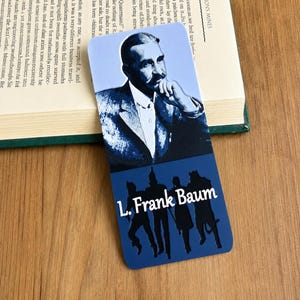 May include: A rectangular bookmark featuring a portrait of L. Frank Baum in shades of blue and white on the top half. The bottom half displays a dark blue background with black silhouettes of four characters and the white text L. Frank Baum.