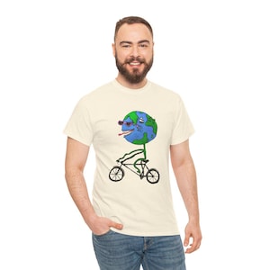 Puede incluir: Camiseta color crema con un personaje de la Tierra de dibujos animados con gafas de sol y sacando la lengua, montando en bicicleta. La Tierra es azul y verde, con un tallo y una bicicleta verdes. La camiseta la lleva una persona.