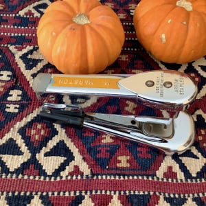 May include: Two orange pumpkins rest on a patterned rug, with a chrome stapler in the foreground. The stapler has a black handle and a yellow label that reads "M. MARSTON". The stapler also has the text "ARROW STAPLES" and "USE ONLY GENUINE NO. 105".