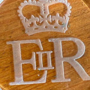 May include: A clear glass coaster with a crown and the letters "ER" etched into the surface. The coaster is set on a wooden surface. The crown is detailed with intricate cutouts.