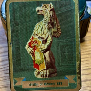 May include: A rectangular tin featuring a detailed illustration of a griffin holding a shield with a crown. The griffin is depicted in shades of beige and brown against a green background. The text "Griffin of Edward III" is displayed on a banner.