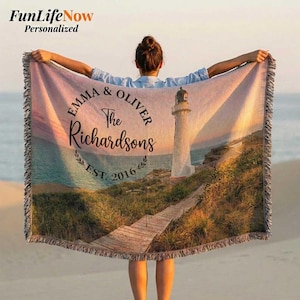 Puede incluir: Una manta tejida personalizada con una escena costera con un faro, una pasarela de madera y vista al mar. La manta tiene un borde con flecos e incluye el texto "Emma & Oliver The Richardsons EST. 2016".