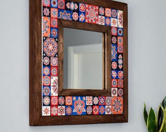 May include: A square mirror with a dark brown wooden frame. The frame is adorned with colourful square tiles displaying detailed floral and geometric designs in red, blue, orange, and white. The mirror reflects a room.
