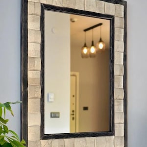 May include: Rectangular wall mirror with a dark wood frame and a decorative border of square, off-white tiles. The mirror reflects a room with hanging lights and a doorway. The mirror is mounted on a light gray wall.