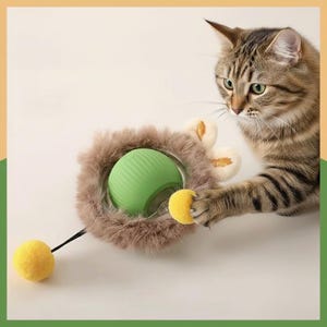 May include: A tabby cat plays with a cat toy. The toy has a green ball in a fluffy, brown ring, with a yellow pom-pom attached. The cat's paw is touching the yellow pom-pom. The background is a light beige colour.