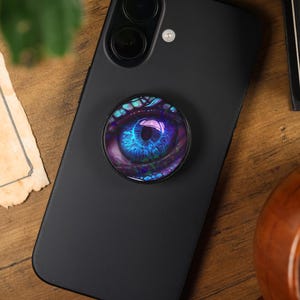 Könnte beinhalten: Ein schwarzes Smartphone mit einem Pop-Socket mit einem detaillierten Augendesign in Blau-, Lila- und Türkistönen. Das Telefon liegt auf einer Holzoberfläche, mit zerrissenem Papier und einer Holztasse im Hintergrund. Der Pop-Socket ist rund.