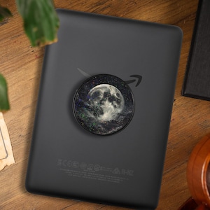 May include: A black e-reader with a round pop socket featuring a full moon and starry night design. The e-reader is on a wooden surface, with a coffee mug and a piece of paper in the background.