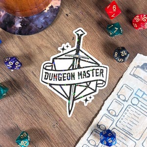 Puede incluir: Una pegatina con el texto "DUNGEON MASTER" y un diseño de espada, rodeada de dados poliédricos rojos, verdes, azules y negros. También se ve una hoja de personaje y una taza de madera.