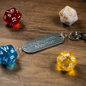 May include: A silver-toned metal keychain tag engraved with the text Dungeon Master, featuring a lobster clasp and a translucent blue twenty-sided die attached to one end, surrounded by a red, a white, and a yellow twenty-sided die.