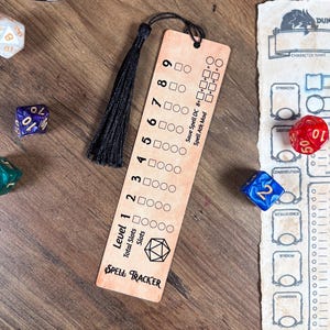 May include: A wooden Spell Tracker bookmark with a black tassel, dice, and a character sheet. The bookmark has a level tracker, spell slots, and text that reads "Spell Tracker". Various multi-sided dice are scattered around the bookmark.