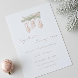 Peut inclure: Une invitation à une baby shower avec le texte "Tiny Booties Coming Soon". L'invitation présente des illustrations à l'aquarelle de chaussons et d'ornements de bébé. Une boule de Noël rose et des branches de pin givrées sont également visibles.
