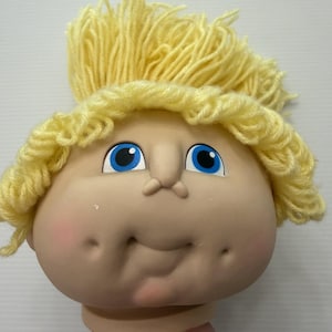 Puede incluir: Una cabeza de muñeca con rostro de vinilo de tono claro, ojos azul brillante, nariz pequeña y mejillas con hoyuelos. La muñeca tiene cabello grueso de lana amarilla texturizada, peinado en capas que enmarcan el rostro de forma definida.