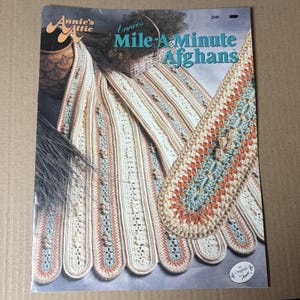 May include: A craft pattern booklet titled Annie's Attic Mile-A-Minute Afghans, featuring a cover image of several long, crocheted strips in cream, light blue, and peach colors with textured bobble stitches and openwork details.