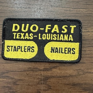 Puede incluir: Un parche bordado rectangular con fondo negro y texto amarillo. El texto dice DUO-FAST TEXAS-LOUISIANA con STAPLERS y NAILERS dentro de dos formas rectangulares redondeadas amarillas. El parche tiene un borde cosido en negro.
