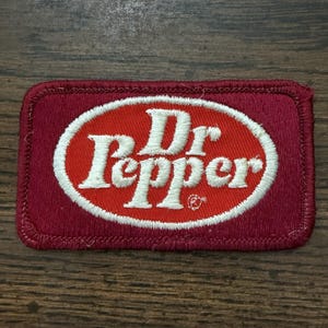May include: A rectangular embroidered patch featuring a maroon background with a central red oval bordered in white. The white embroidered text reads Dr Pepper in a stylized script font, with a small registered trademark symbol near the letter r.