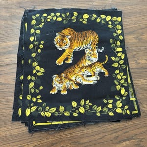 May include: A stack of square fabric panels featuring a black background with a central illustration of three tigers in orange, black, and white. The design is framed by a border of yellow and green leaves, with frayed edges on the fabric squares.