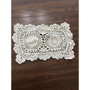 May include: A rectangular off-white crochet doily featuring two circular motifs connected by a central pattern. The piece has a scalloped, decorative border with intricate openwork stitching throughout, creating a detailed, textured textile design.