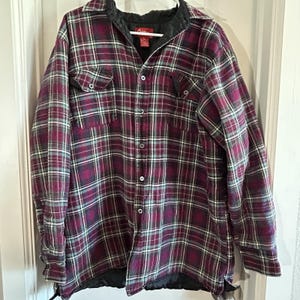 May include: A burgundy, teal, and white plaid flannel shirt with a button-down front. The shirt features two chest pockets and a black quilted lining. The collar is black, and the shirt is hanging on a white hanger.