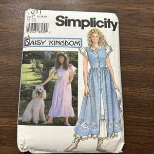 May include: A Simplicity Daisy Kingdom sewing pattern envelope for a dress in size P 12, 14, 16. The front features an illustration of a blue plaid dress with ruffled sleeves and a white floral border, alongside a photograph of a person with a poodle.
