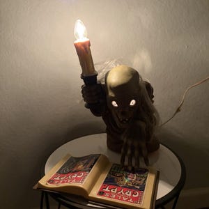 May include: A light-up figurine of a skeletal figure with white hair holding a lit candle, positioned behind an open book with Tales from the Crypt comic book covers printed on the pages. The figure has glowing white eyes and a reaching hand.