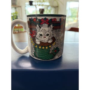 May include: A white ceramic mug featuring a graphic illustration of a fluffy grey kitten peeking out of a green Christmas stocking. The stocking is decorated with small yellow bells and a red ribbon, set against a stone fireplace background.