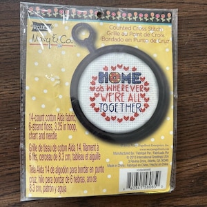 May include: A counted cross-stitch kit featuring a design with the word "HOME" and the phrase "WHEREVER WE'RE ALL TOGETHER" in a heart shape. Includes 14-count Aida fabric, 3.25-inch hoop, floss, chart, and needle.