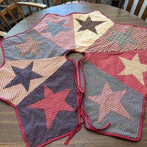 May include: A circular fabric tree skirt featuring a patchwork design of alternating triangular panels. The panels display various patterns including red, blue, and black gingham, stripes, and solid colors, each decorated with a large fabric star.