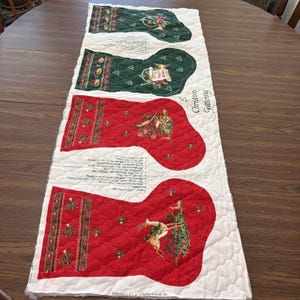 May include: A quilted table runner featuring four printed Christmas stockings in alternating red and green colors on a cream background. The design includes illustrations of deer, trumpets, and holly, with printed text reading A Christmas Gathering.