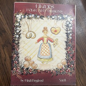 May include: A softcover book titled Heidi's Folk Art Designs Vol. II by Heidi England. The cover features a central illustration of a person in a dress, a heart, and a circular ornament, surrounded by a border of holly leaves, berries, and pearls.