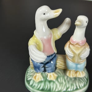 May include: A ceramic figurine featuring two anthropomorphic ducks. One duck is gesturing with its wing, wearing a yellow jacket, pink shirt, and blue pants. The other duck wears a yellow vest, blue shorts, and a pink bow tie. The base is green with grass.