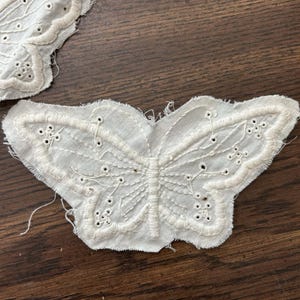 May include: Two white, embroidered butterfly appliques with detailed wing patterns. The butterflies are made of a textured fabric with delicate stitching and small cut-out details. The appliques are placed on a dark wood surface.