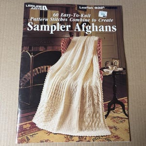 May include: A Leisure Arts knitting pattern booklet titled 60 Easy-To-Knit Pattern Stitches Combine to Create Sampler Afghans, featuring a cream-colored knitted blanket with various textured stitch patterns draped over a chair.