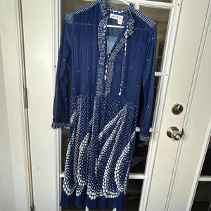 Puede incluir: Vestido azul marino vintage con lunares blancos y patrones de ondas. El vestido presenta mangas largas, una falda plisada y un cuello alto con corbata. El vestido está colgado en una percha blanca.