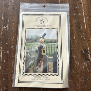 May include: A sewing pattern packet titled Percy Pumpkin 23.5" by Kimberly R. Whitner. The cover features a photograph of a fabric doll with a pumpkin head, wearing a black top hat, tan shirt, and plaid pants, holding a small black cat figure.