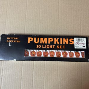 May include: A rectangular black box labeled BATTERY OPERATED PUMPKINS 10 LIGHT SET. The packaging features a clear window displaying ten small, orange, translucent pumpkin-shaped light covers with carved jack-o'-lantern faces.