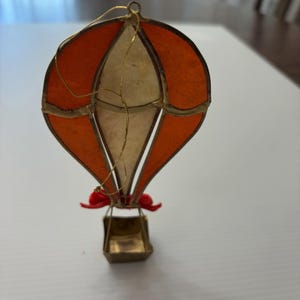 May include: A hot air balloon ornament featuring orange and off-white translucent panels framed in gold-toned metal. The structure includes a small gold-toned metal basket at the base, accented with two small red fabric bows and a gold hanging string.