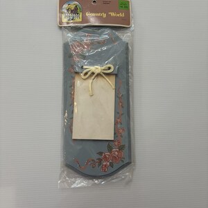 May include: A blue wooden wall plaque in clear plastic packaging with a Country World header card. The plaque features painted pink roses and green leaves, with a cream-colored rectangular paper notepad attached by a cream-colored string bow.