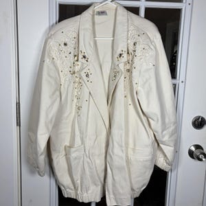 May include: An off-white jacket featuring long sleeves, a collared neckline, and two front pockets. The upper chest area is decorated with intricate white embroidery, small metallic studs, and clear faceted beads arranged in a symmetrical pattern.