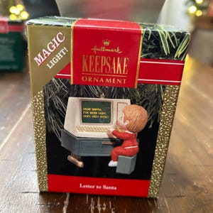 Könnte beinhalten: Hallmark Keepsake Weihnachtsschmuck in einer Box. Der Schmuck zeigt ein Kind in einem roten Anzug, das auf einem Computer tippt, mit der Nachricht "Lieber Weihnachtsmann, ich war sehr, sehr, sehr brav!" Die Box hat goldene Glitzer und rote Akzente.
