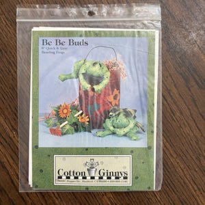 May include: A craft pattern packet titled Be Be Buds 8" Quick & Easy Beanbag Frogs by Cotton Ginnys. The cover features two green fabric frogs, one inside a patterned container and one sitting beside it with artificial flowers and greenery.