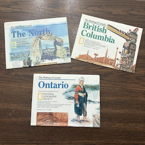Puede incluir: Tres suplementos de mapas de la National Geographic Society titulados The Making of Canada. Los mapas muestran The North con un inukshuk, British Columbia con un tótem y Ontario con una figura histórica frente a las cataratas del Niágara.