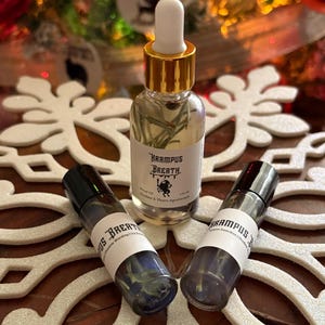 May include: A clear glass bottle with a gold dropper top and a label that reads "Krampus Breath". Two roller bottles with the same label are also present. The bottles are on a white snowflake-shaped decoration.