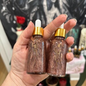 May include: Two clear glass dropper bottles filled with a shimmering, metallic bronze liquid containing small, thin, light-colored botanical pieces. Each bottle features a gold-colored collar and a white rubber dropper bulb top.