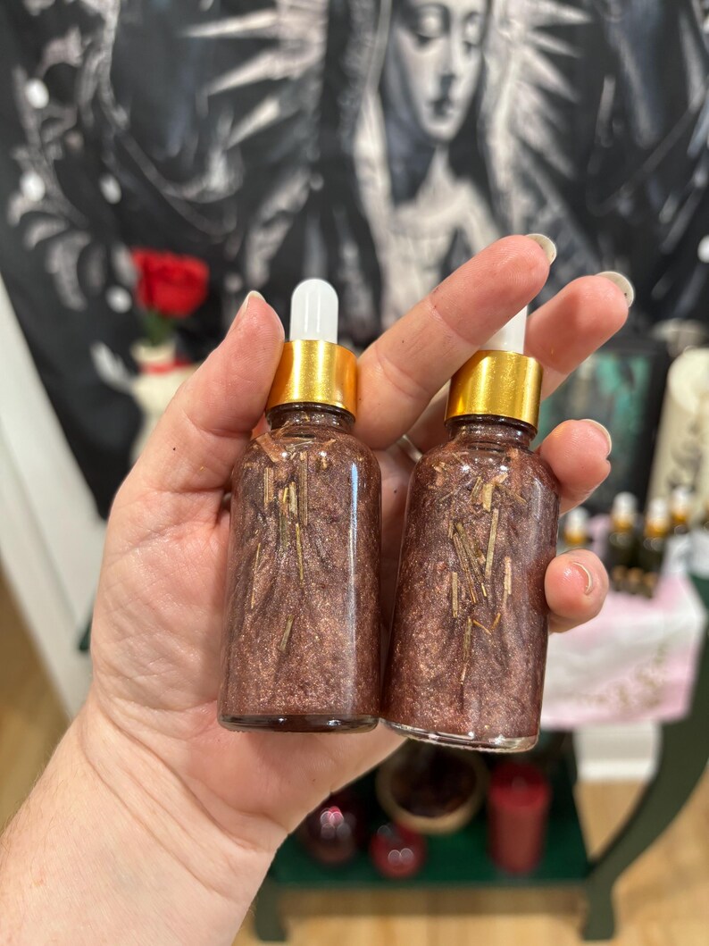 May include: Two clear glass dropper bottles filled with a shimmering, metallic bronze liquid containing small, thin, light-colored botanical pieces. Each bottle features a gold-colored collar and a white rubber dropper bulb top.